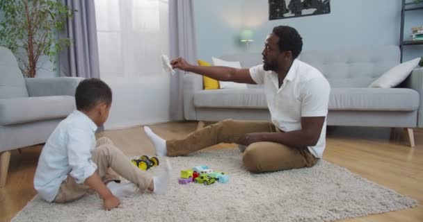 Niño alegre jugando con el padre mientras está sentado en el suelo en la sala de estar. alegre afroamericano guapo hombre divertirse con lindo hijo y jugar con avión juguete en casa. Concepto de infancia — Vídeos de Stock