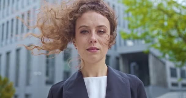 Portrét dobře vypadající kudrnaté ženy ve společenském obleku venku. Sebevědomá běloška, která stojí poblíž moderní budovy a dívá se do kamery. Obchod, koncepce řízení. — Stock video