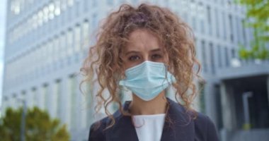 Açık havada resmi takım elbiseli çekici kıvırcık saçlı kadın çalışan. Sağlık maskesi takmış beyaz bir iş kadını kameraya bakıyor. Koruma, sağlık, koronavirüs..