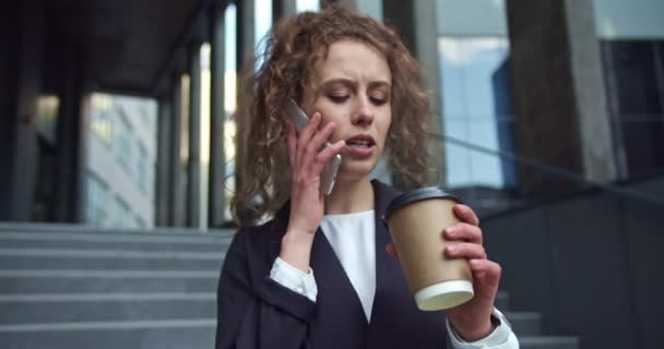 Confident Caucasian businesswoman wearing a formal suit with a smartwatch in business district. Beautiful female office worker walking near modern building, drinking coffee and talking on the phone. — Stock Video