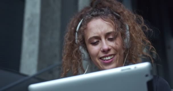 Pretty Caucasian business lady wearing a formal suit, in headphones near the modern buildings. Smiling office worker scrolling the news feed and listening to music on the tablet. Wellness lifestyle. — Stock Video