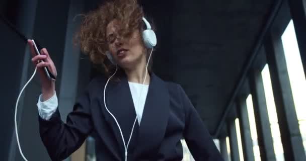 Joyful businesswoman in a formal suit and headphones going down the stairs. Pretty curly haired female worker dancing and listening to music on the smartphone. Modern buildings background. — Stock Video