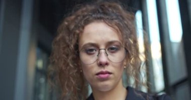 Beyaz tenli, gözlüklü, resmi takım elbiseli bir finans işçisi. Güzel kıvırcık saçlı iş kadını kameraya bakıyor ve göz kırpıyor. Modern bölge geçmişi.