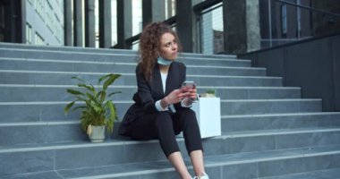 Üzgün, modern binanın dışında resmi bir maske takan beyaz bir kadını kovdu. Güzel kıvırcık saçlı kadın merdivenlerde oturuyor, yeşillik içinde bir bitki ve bir de iş eşyası var. Görevden alınma.