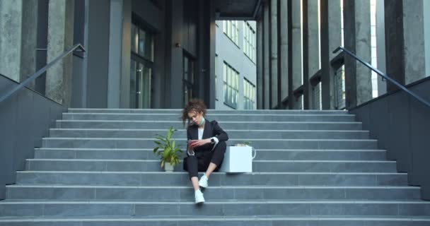 Rozrušená běloška v obleku a lékařské masce s krabicí a rostlinou. Propuštěný kancelářský pracovník je smutný, sedí na schodech poblíž budovy společnosti a používá smartphone. Propuštění. — Stock video