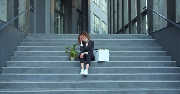 Elle a viré une employée de bureau caucasienne portant un costume près du bâtiment moderne. Femme d'affaires frustrée assise dans les escaliers avec une boîte et une plante, grisonnante. Cessation d'emploi, concept de chômage. — Video