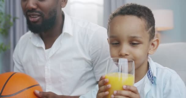 Primer plano retrato de lindo niño afroamericano feliz beber jugo mientras está sentado en casa con el padre. Pequeño niño alegre adorable degustación de jugo de buen humor en la sala de estar. Concepto de infancia — Vídeos de Stock