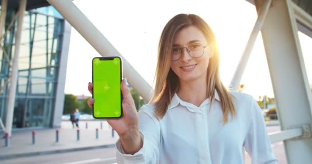 Lviv, Ucrania, 5.08.2019: Retrato de una chica bonita y feliz con gafas de pie en la ciudad y mostrando el teléfono inteligente con pantalla verde con movimiento de seguimiento. Hermosa mujer sosteniendo el teléfono celular con croma key — Vídeos de Stock