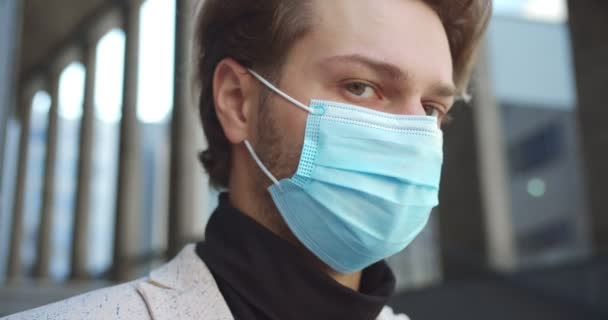 Primer plano retrato de hombre guapo trabajador en máscara médica de pie al aire libre en la calle y mirando a la cámara. Hombre serio caucásico en cuarentena. Protección contra virus en la ciudad. Concepto de trabajo — Vídeos de Stock