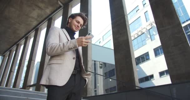 Dolny widok szczęśliwy biały mężczyzna profesjonalne pisanie i stukanie na smartfonie podczas stania na świeżym powietrzu. Przystojny, radosny młodzieniec, piszący na komórkę na ulicy i uśmiechający się. Koncepcja przedsiębiorstwa — Wideo stockowe