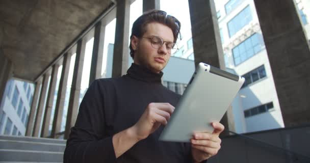 Retrato de guapo gerente masculino serio escribiendo en la tableta mientras está parado al aire libre solo. Caucásico concentrado joven empresario golpeando y navegando en el dispositivo en la calle. Concepto de trabajo — Vídeo de stock