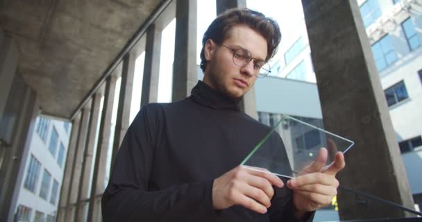 Retrato de belo trabalhador masculino sério em óculos deslizando em gadget futurista enquanto está ao ar livre. Caucasiano concentrou jovem empresário usando holograma dispositivo na rua. Conceito de tecnologia — Vídeo de Stock