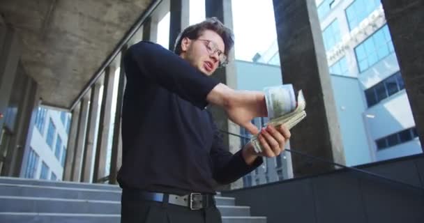 Retrato de feliz divertido hombre de negocios caucásico dispersando dólares mientras está parado solo en las escaleras al aire libre. Un joven guapo que hace llover dinero tirando dinero al aire en la ciudad. Concepto de dinero — Vídeo de stock