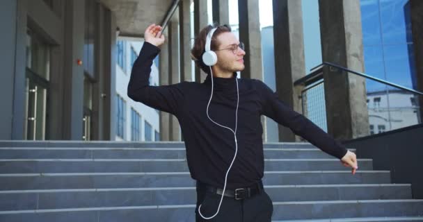 Portret van een gelukkige jongeman die naar muziek luistert in een koptelefoon en danst op een trap buiten in de stad. Knappe blanke vrolijke man met bril luistert naar liedjes en beweegt zich op straat. Vrijetijdsconcept — Stockvideo