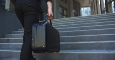 Ofis binasının yanında üst katta yürürken beyaz bir erkeğin siyah evrak çantası tutarken yakın plan çekimleri. Şehirde çantalı bir adam var. Ofis çalışanı. İş konsepti