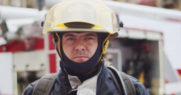 Close up portrait of caucasian handsome fireman in helmet and gull equipment standing next to the car and looking into camera. The concept of saving lives, heroic profession, fire safety — Stock Video