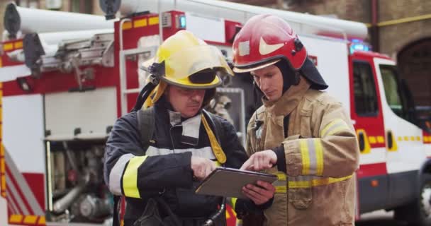Plano medio de dos bomberos discutiendo plan de rescate mirando la tableta mientras están de pie en cascos cerca de una furgoneta de bomberos. El concepto de salvar vidas, profesión heroica, seguridad contra incendios — Vídeos de Stock