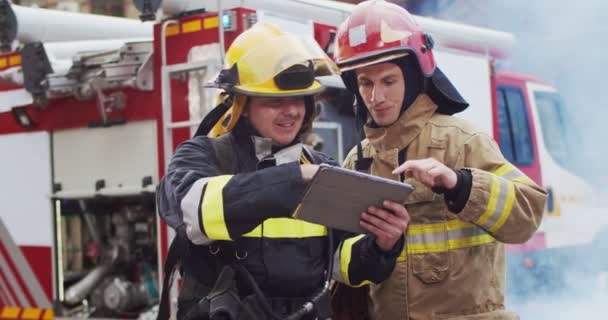 Střední plán dvou hasičů diskutujících o záchranném plánu při pohledu na tablet v helmách poblíž hasičské dodávky. Kouř z požárních krytů zachránců a hasičské auto. — Stock video