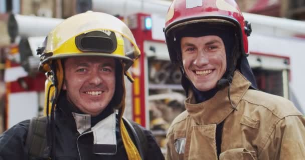 Ritratto ravvicinato di due vigili del fuoco in casco e attrezzature gabbiano in piedi accanto alla macchina e sorridente guardando la fotocamera. Concetto di salvare vite umane, professione eroica, sicurezza antincendio — Video Stock