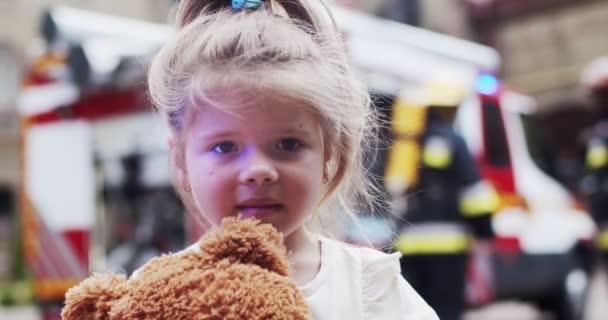 Close up portrait of little cute girl with teddy bear standing near firefighters with a fire truck after rescue and looking at camera. The concept of saving lives, heroic profession, fire safety — Stock Video