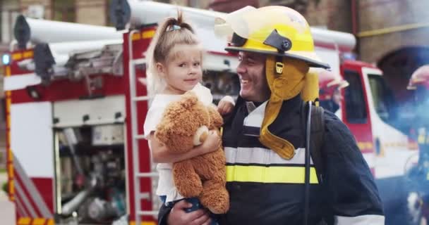 Portrait of caucasian handsome fireman in helmet and gull equipment holds girl with teddy bear in arms and looking into camera smiling. The concept of saving lives, heroic profession, fire safety — Stock Video