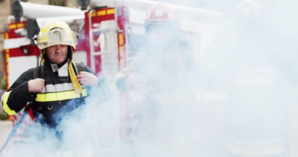 Piano intermedio di tre vigili del fuoco in casco e attrezzature gabbiano in piedi accanto alla macchina e guardando la fotocamera. Il fumo del fuoco copre i soccorritori. Concetto di salvare vite umane, professione eroica, sicurezza antincendio — Video Stock