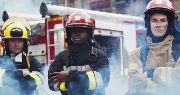 Крупный план трех пожарных в шлеме и чайка оборудования, стоящих рядом с автомобилем и глядя на камеру. Дым от огня прикрывает спасателей. Концепция спасения жизней, героической профессии, пожарной безопасности — стоковое видео