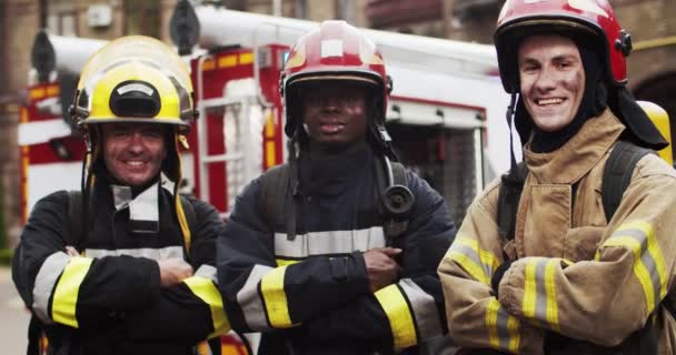 Portrait of three happy satisfied firefighters standing next to fire van in front of camera looking and smiling crossing hands after hard work. Concept of saving lives, heroic profession, fire safety — Stock Video