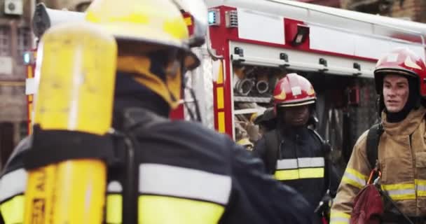 Tres bomberos de diferentes razas en cascos y equipo de gaviotas discutiendo plan de rescate de pie cerca de furgoneta de bomberos. Bombero con tableta en las manos mostrando dirección. Humo de salvadores de cubiertas contra incendios. — Vídeo de stock