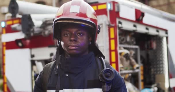 Портрет молодого радостного симпатичного афроамериканского пожарного в шлеме, смотрящего в камеру рядом с пожарной машиной. Концепция спасения жизней, героической профессии, пожарной безопасности — стоковое видео
