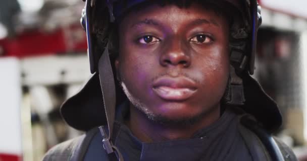 Zblízka portrét afroamerického fireguardu v helmě hledící do kamery vedle hasičského vozu s blikajícími světly. Koncept záchrany životů, hrdinské profese, požární bezpečnosti — Stock video