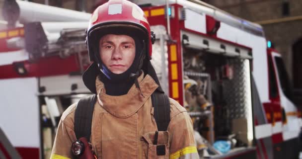 Plano medio de bombero guapo caucásico en casco y equipo de gaviota de pie junto al coche con luces intermitentes encendidas y mirando a la cámara. El concepto de salvar vidas, profesión heroica, seguridad contra incendios — Vídeos de Stock