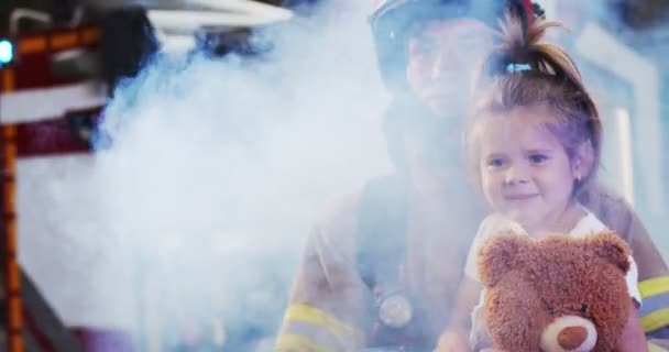 Portrait of firefighter approaches from fire van with cute girl on arms and flashing lights on and standing in front of camera. Smoke from fire covers rescuers. Concept of saving lives, fire safety — Stock Video