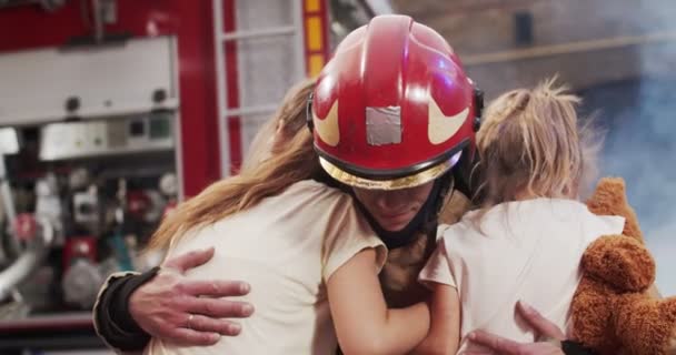 Portrait of injured little girls sisters reunites with their loving father fireman in helmet and gull equipment. Smoke from fire covers rescuers and car. Firefighter looking at camera and smiling. — Stock Video