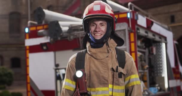 Plano medio de bombero guapo caucásico en casco y equipo de gaviota brazos cruzados de pie junto al coche y felizmente mirando a la cámara sonriendo. Concepto de salvar vidas, profesión heroica, seguridad contra incendios — Vídeos de Stock