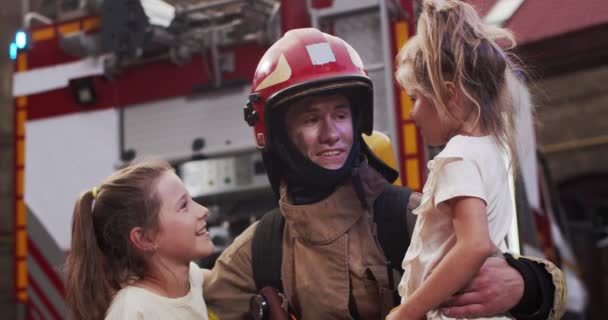 Ritratto del bel giovane vigile del fuoco caucasico che tiene in mano due piccole ragazze salvate mentre guardano la macchina fotografica e sorridono felici mentre sono all'aperto accanto al grande sfondo rosso del camion dei pompieri. — Video Stock