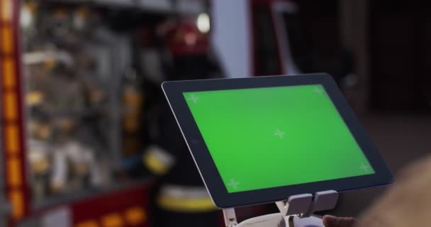 Крупним планом пожежник дивиться на масштаб вогню, використовуючи екран, що транслюється з безпілотника. Зелений екран на планшеті на фоні пожежного відділення. Концепція порятунку життя, героїчна професія, пожежна безпека — стокове відео