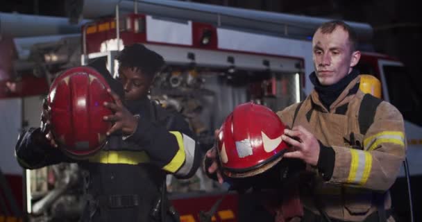 Plano medio de dos bomberos afroamericanos y caucásicos en equipo de gaviota de pie junto al coche y quitándose el casco. Concepto de salvar vidas, profesión heroica, seguridad contra incendios — Vídeos de Stock