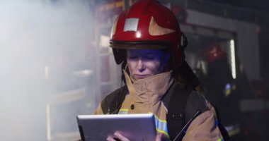Yetişkin beyaz kadın itfaiyecinin portresi. Tablet cihazına bakıyor. Gece duman içinde itfaiye arabasının yanında duruyor. Hayat kurtarma, kahramanlık mesleği, yangın güvenliği kavramı.