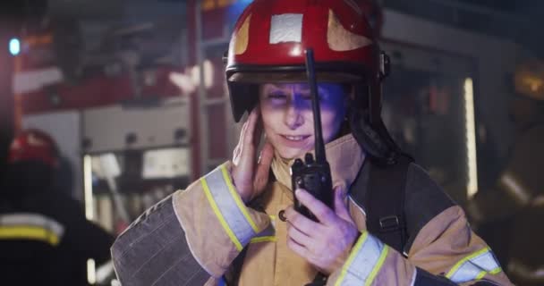 Retrato de una bombera confiada en casco y equipo de gaviota parada junto al coche con luces intermitentes encendidas por la noche explicando el despachador en walkie-talkie. Concepto de salvar vidas, seguridad contra incendios — Vídeo de stock