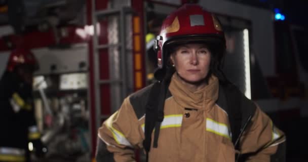 Retrato de bombero femenino se acerca desde la furgoneta de bomberos con luces intermitentes encendidas por la noche y de pie delante de la cámara mirando. El concepto de salvar vidas, profesión heroica, seguridad contra incendios — Vídeo de stock