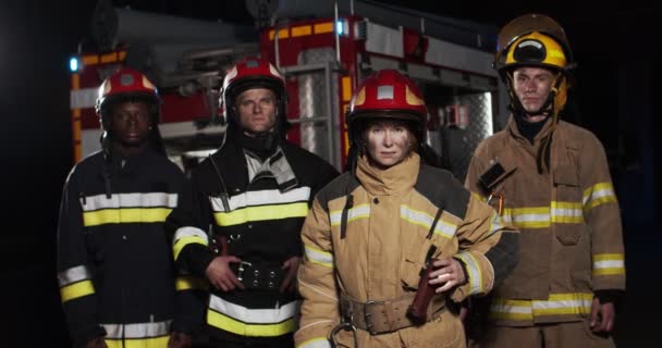 Plano medio de cuatro bomberos que llevan equipo completo con herramientas de rescate de emergencia de pie junto al coche por la noche y mirando a la cámara. Camiones de humo y bomberos en el fondo. — Vídeo de stock
