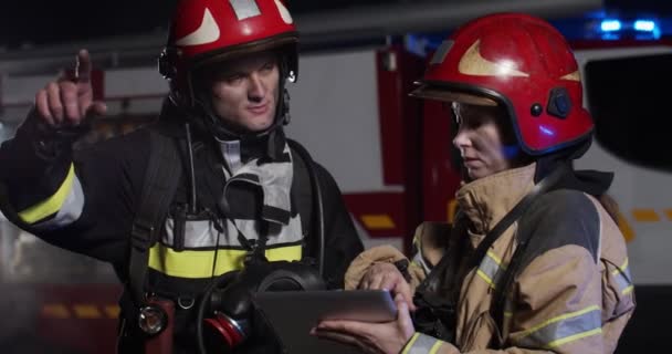 Primer plano de los bomberos caucásicos con los trajes y cascos completamente equipados de pie afuera, mirando algo en la computadora de la tableta y disputando por la noche. — Vídeos de Stock