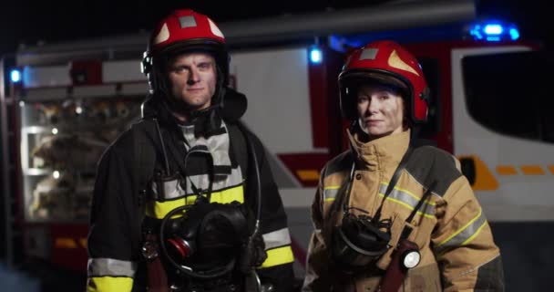 Plano medio de hombres y mujeres bomberos caucásicos con los trajes y cascos completamente equipados de pie afuera mirando a la cámara por la noche. Concepto de salvar vidas, profesión heroica, seguridad contra incendios — Vídeos de Stock