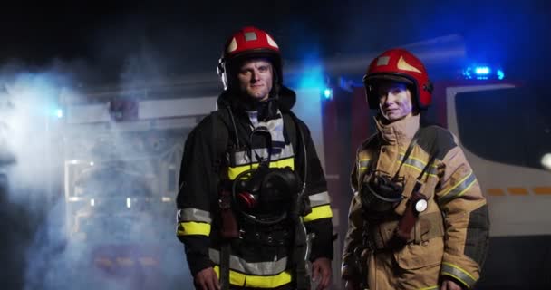 Plano medio de bomberos caucásicos con los trajes y cascos completamente equipados de pie afuera mirando a la cámara por la noche en humo junto a la furgoneta de bomberos con luces intermitentes encendidas. — Vídeo de stock