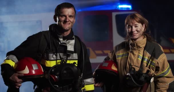 Portret kaukaska kobieta i mężczyzna strażak w mewie sprzęt trzymając kask w rękach patrząc w kamerze i uśmiechając się w pobliżu wozu strażackiego z dymem w nocy. Koncepcja ratowania życia, bezpieczeństwa przeciwpożarowego — Wideo stockowe