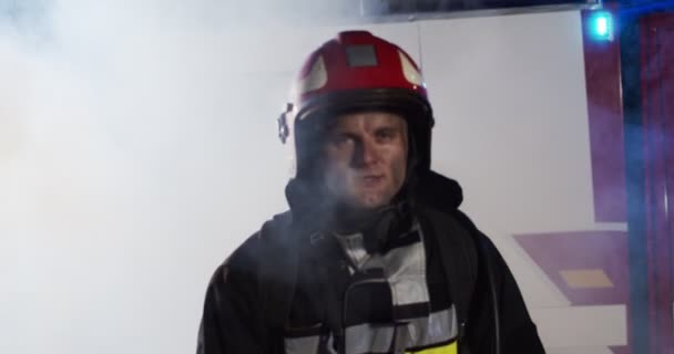 Close up of firefighter approaches from fire van with flashing lights on and standing in front of camera looking and smiling after hard work. Concept of saving lives, heroic profession, fire safety — Stock Video