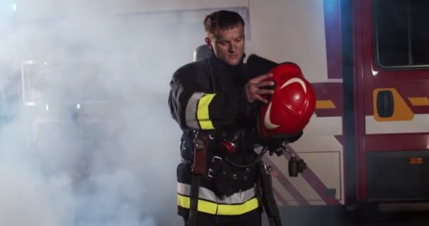 Střední plán unaveného mladého bělocha hasiče, který si sundává helmu, vychází z ohně na červený náklaďák a dívá se do kamery. Koncept záchrany životů, hrdinské profese, požární bezpečnosti — Stock video