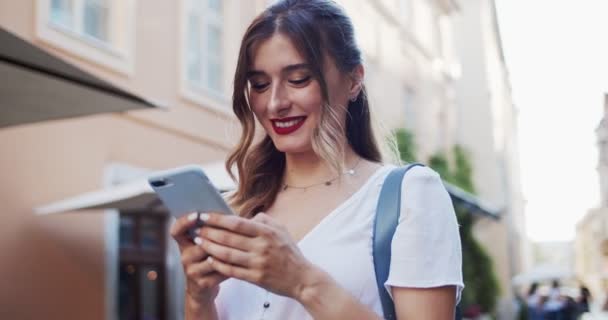 야외를 걷는 동안 모빌 폰을 사용하여 행복 한 젊은 금발 여성의 사진을 클로즈업 한다. 아름다운 소녀는 뉴스를 읽으면서 웃고 시내 거리에서 핸드폰 화면을 보고 있습니다. 실제 생명에 대한 개념 — 비디오