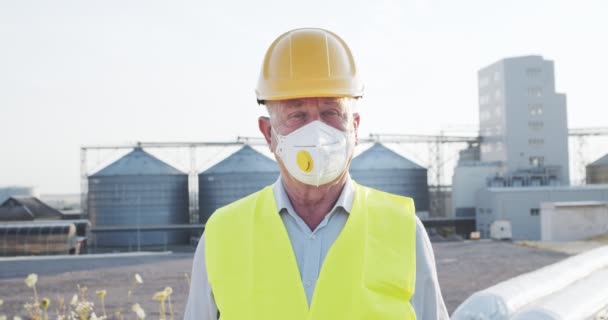 마스크를 쓰고 현실감 있게 헬멧을 쓰고 대형 산업 공장 옆에 서서 햇빛을 받으며 카메라를 보고 있는 산업 고위 기술자의 모습. 산업 및 공학 연구 개념 . — 비디오